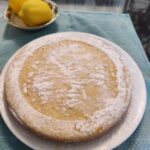 Bowl with 2 lemons next to an 8-inch round Lemon Vanilla Cake lightly dusted with powdered sugar
